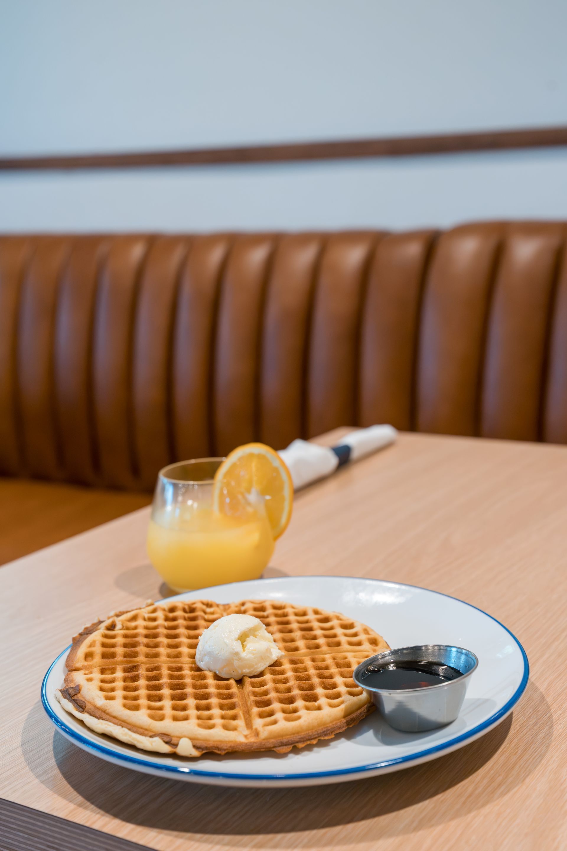Waffle with butter and syrup on a white plate beside a lemon drink on a wooden table, with a brown bench behind