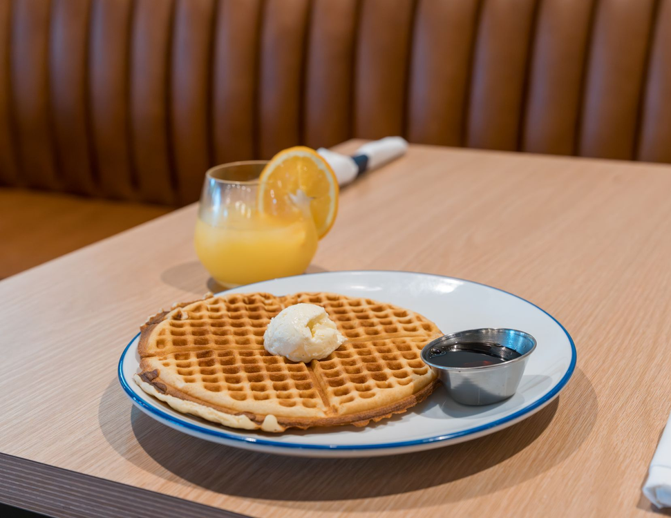 Waffle with butter and syrup on a white plate beside a glass of orange juice on a café table