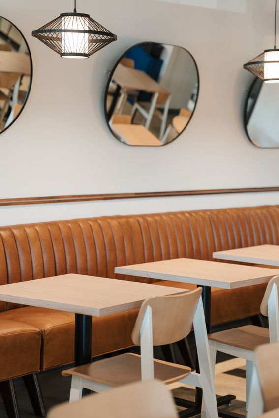 Empty café seating with light wood tables, white chairs, orange banquette, and round mirrors on the wall