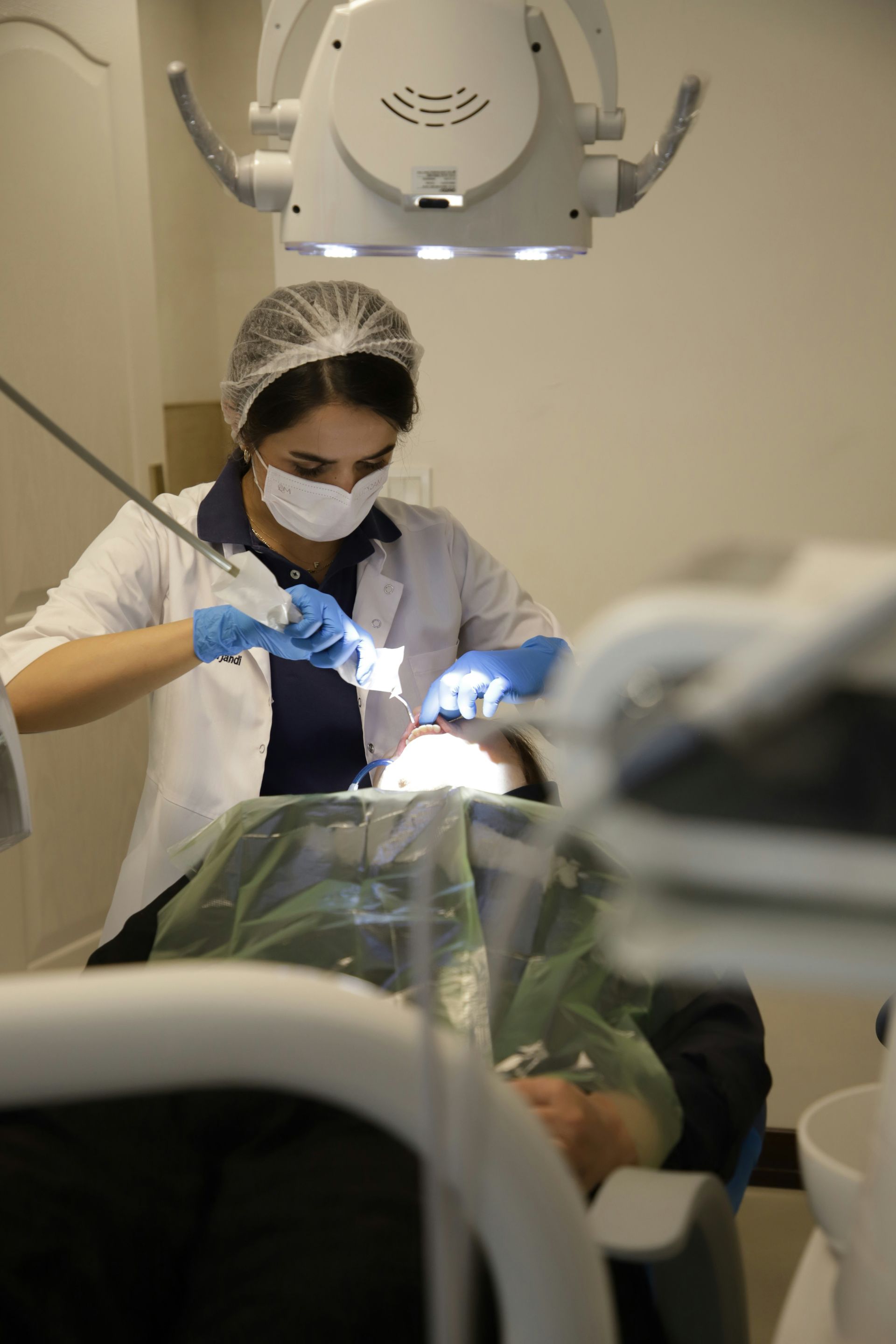 Dentist in white coat, mask, and gloves working on a patient's teeth under bright overhead light.