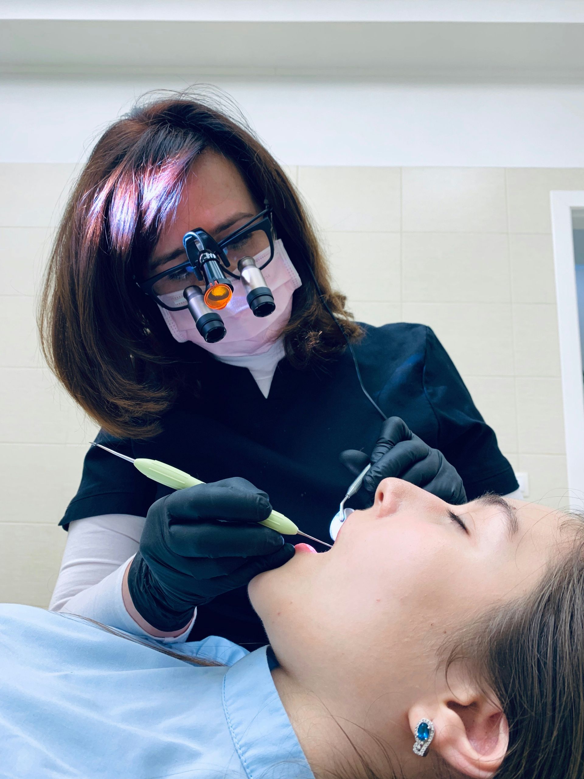 Dentist examining a patient's mouth. Dentist wears a mask, loupes, black gloves and scrubs. Patient is in a dental chair.
