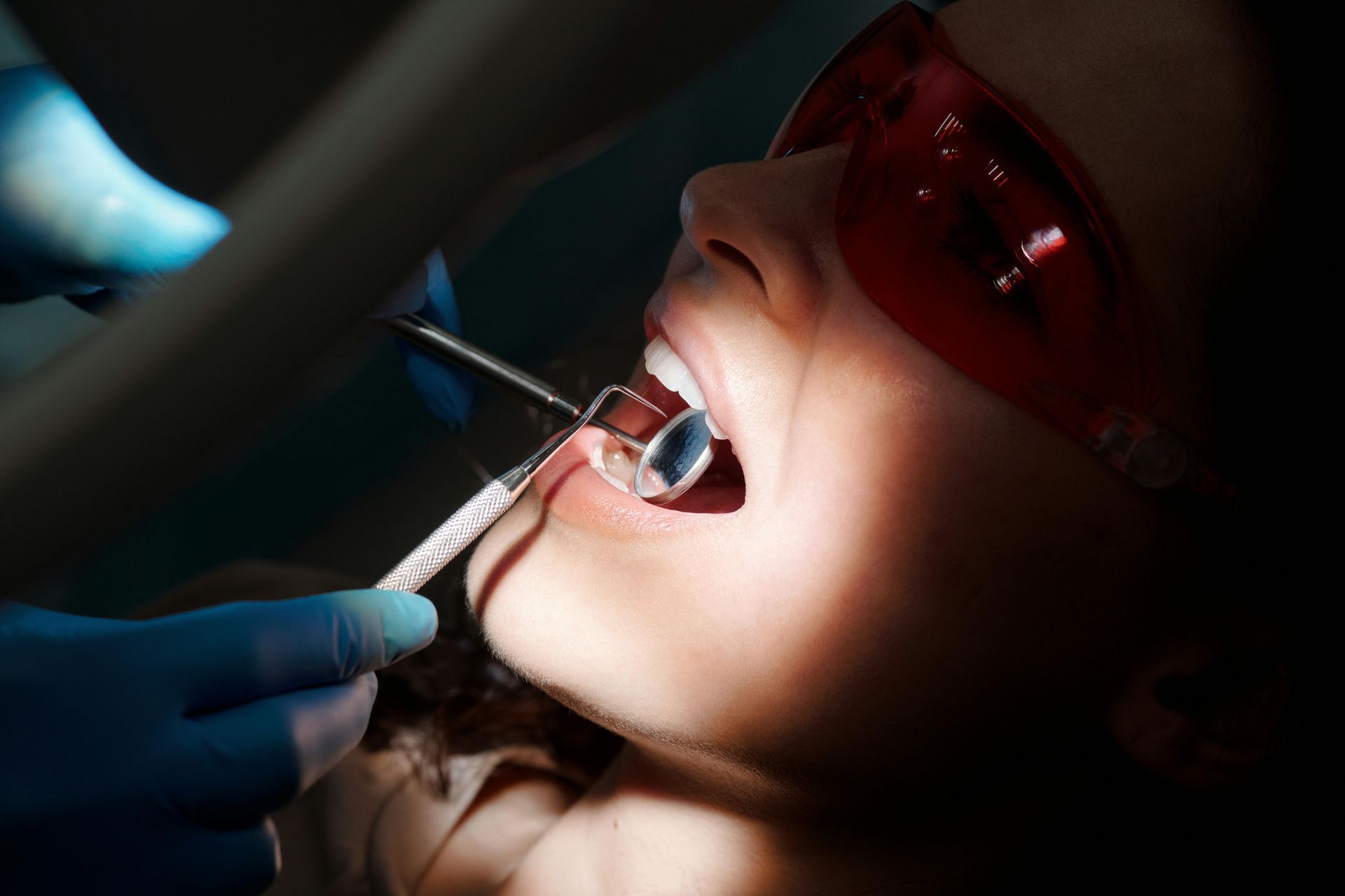 Person in dentist chair, mouth open, being examined with tools. Wearing protective eyewear.