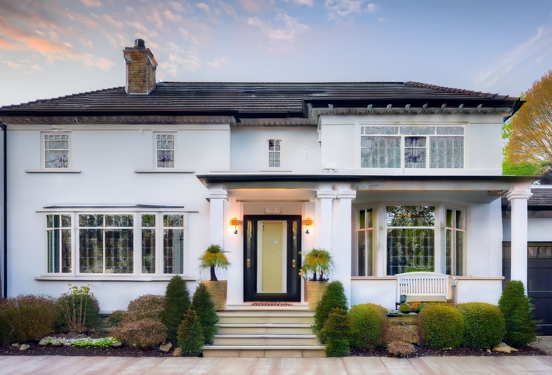 White two-story house with black roof, front steps, large windows, and manicured shrubs at dusk