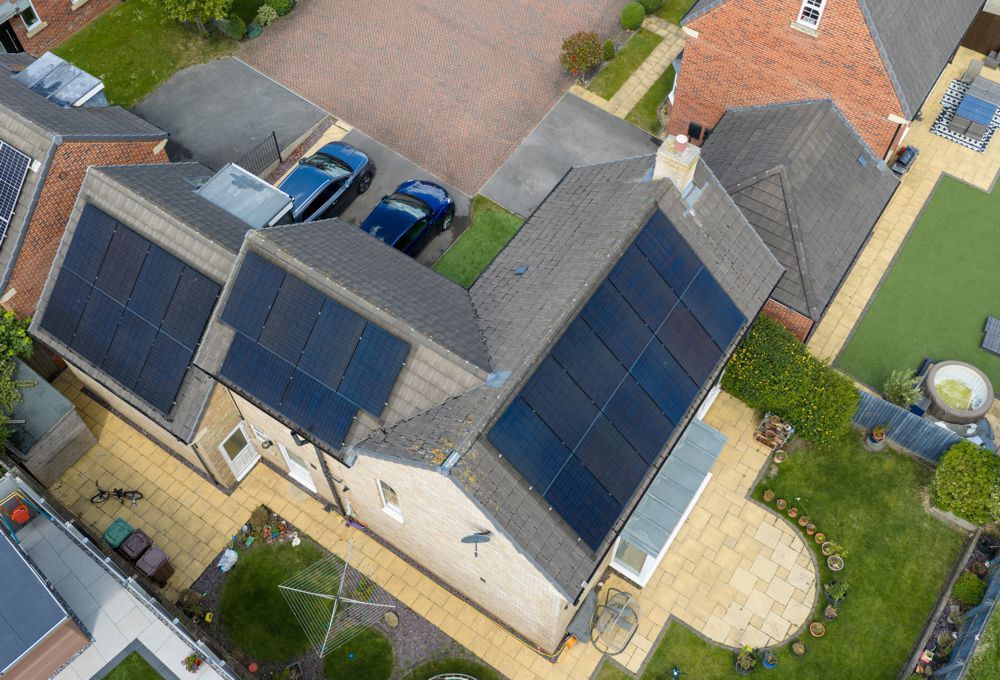 Aerial view of a house with solar panels on two roof sections, driveway, and neighboring homes.