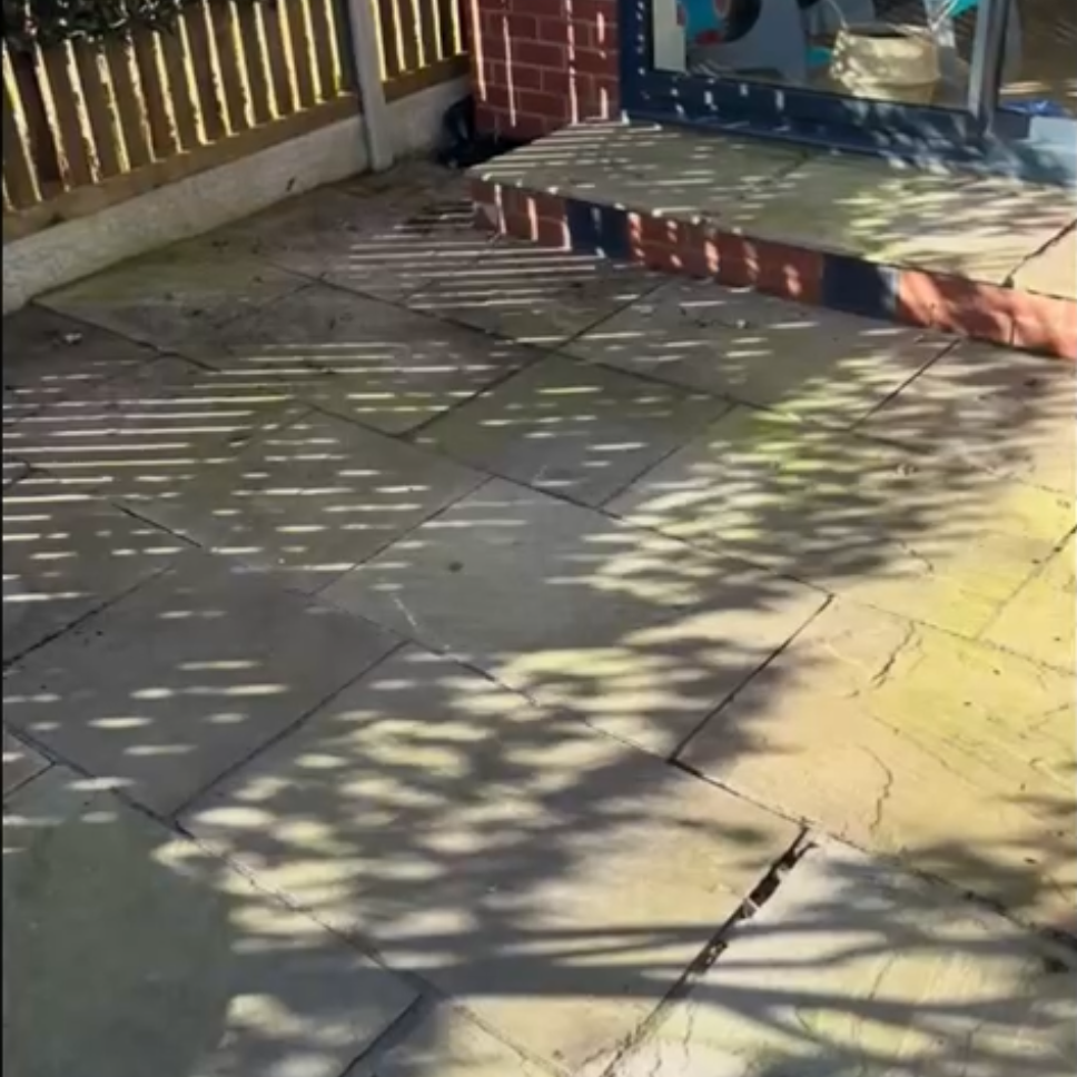 Sunlit backyard patio with large gray paving slabs, steps, and dappled tree shadows.