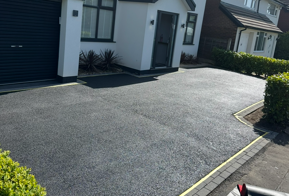 Driveway in front of a white house with a garage and front door, bordered by hedges and paving stones.