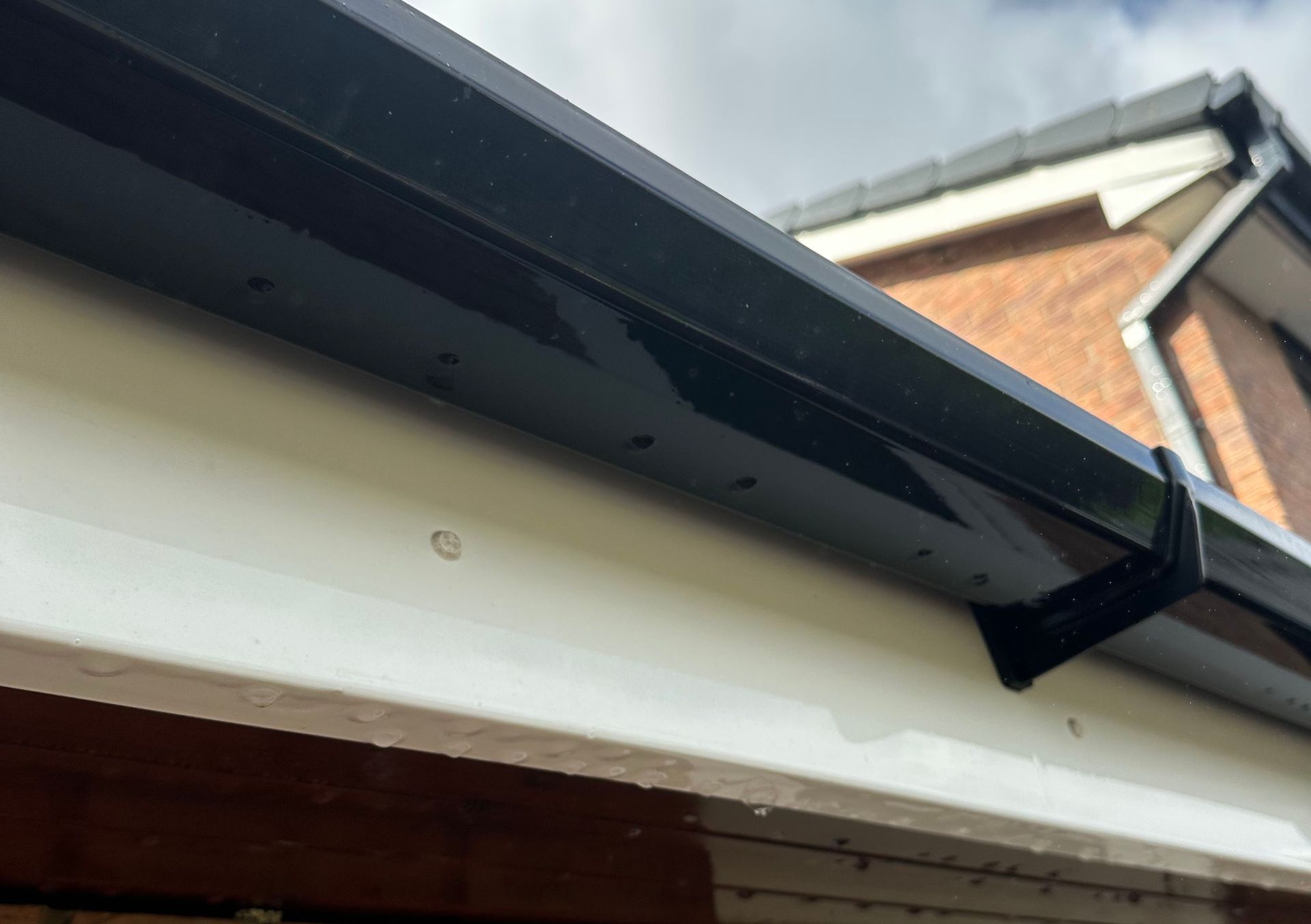 Close-up of a house roof edge with black guttering and white fascia under a brick wall and cloudy sky