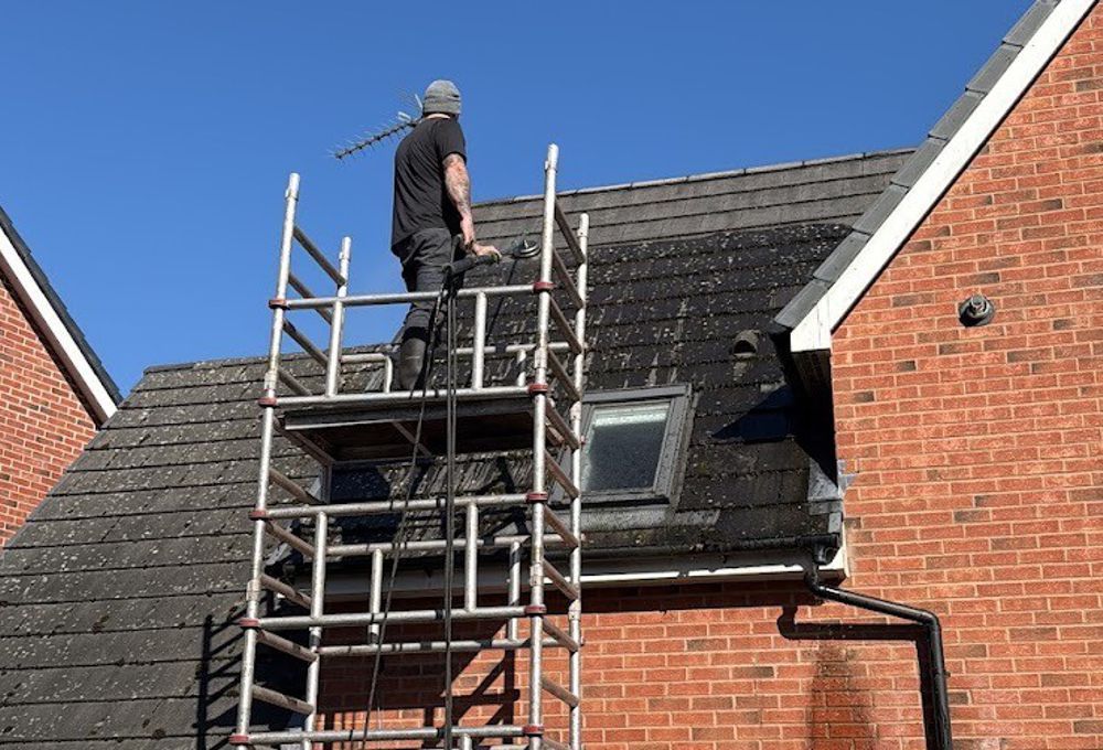 Worker on scaffolding repairing a slate roof beside a brick house under a clear blue sky