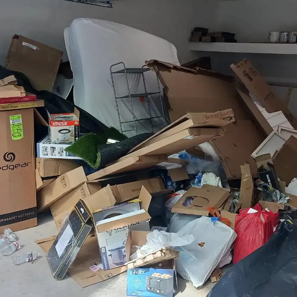 Messy room filled with piled cardboard boxes, trash bags, and clutter around a covered chair
