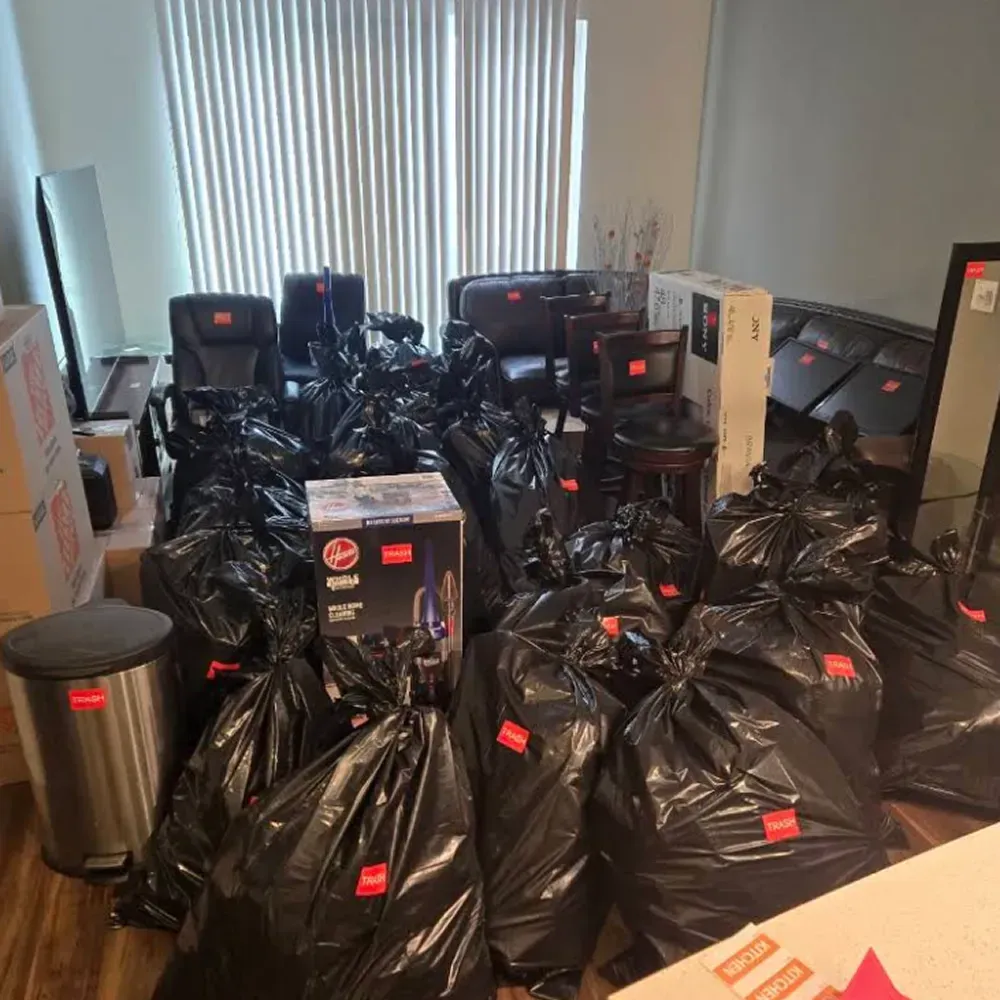 Many black trash bags piled in a room with boxes, monitors, and a metal bin
