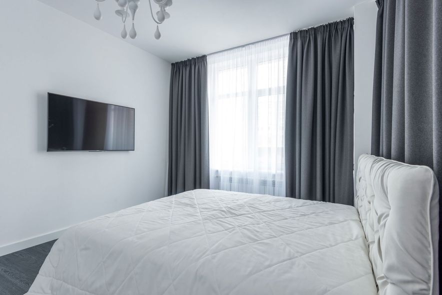 Bright white bedroom with a bed, wall-mounted TV, and tall gray curtains over a window.