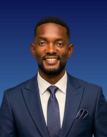 A professional headshot of a person wearing a navy blue suit, white shirt, and patterned tie against a blue background.