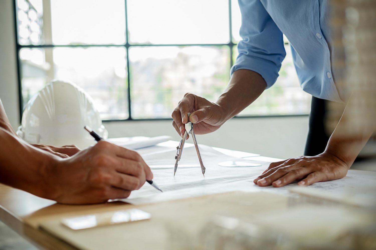 Two people work together at a desk, using a drafting compass and pen on technical blueprints with a white hard hat nearby.