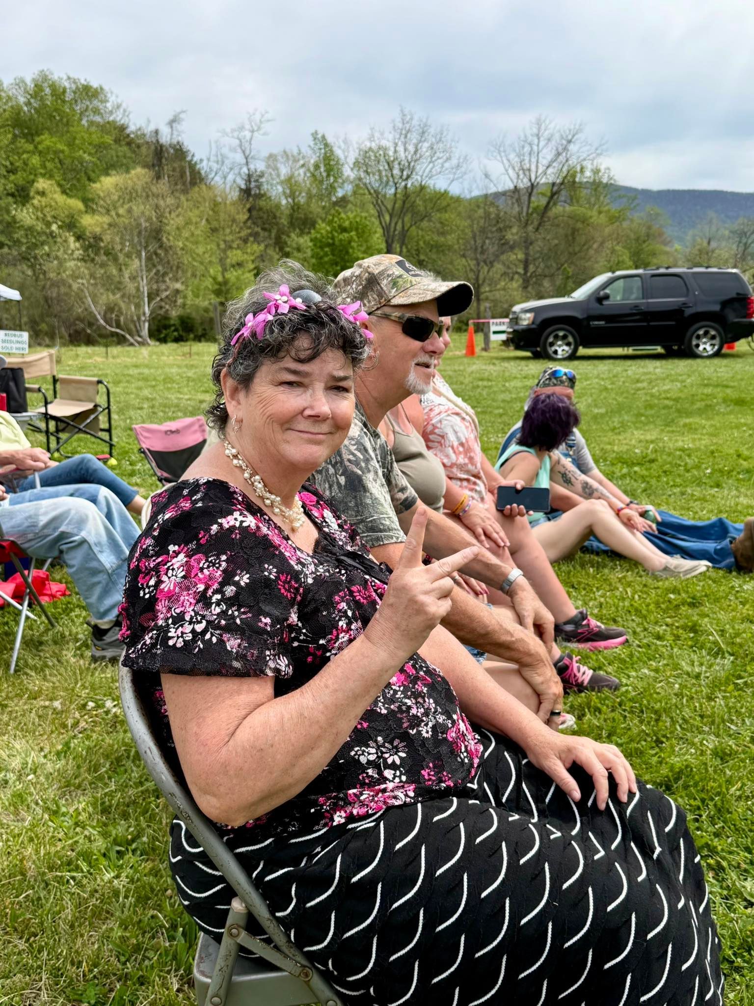 Smiling woman in black floral dress and pink flower crown sits on grass at an outdoor gathering
