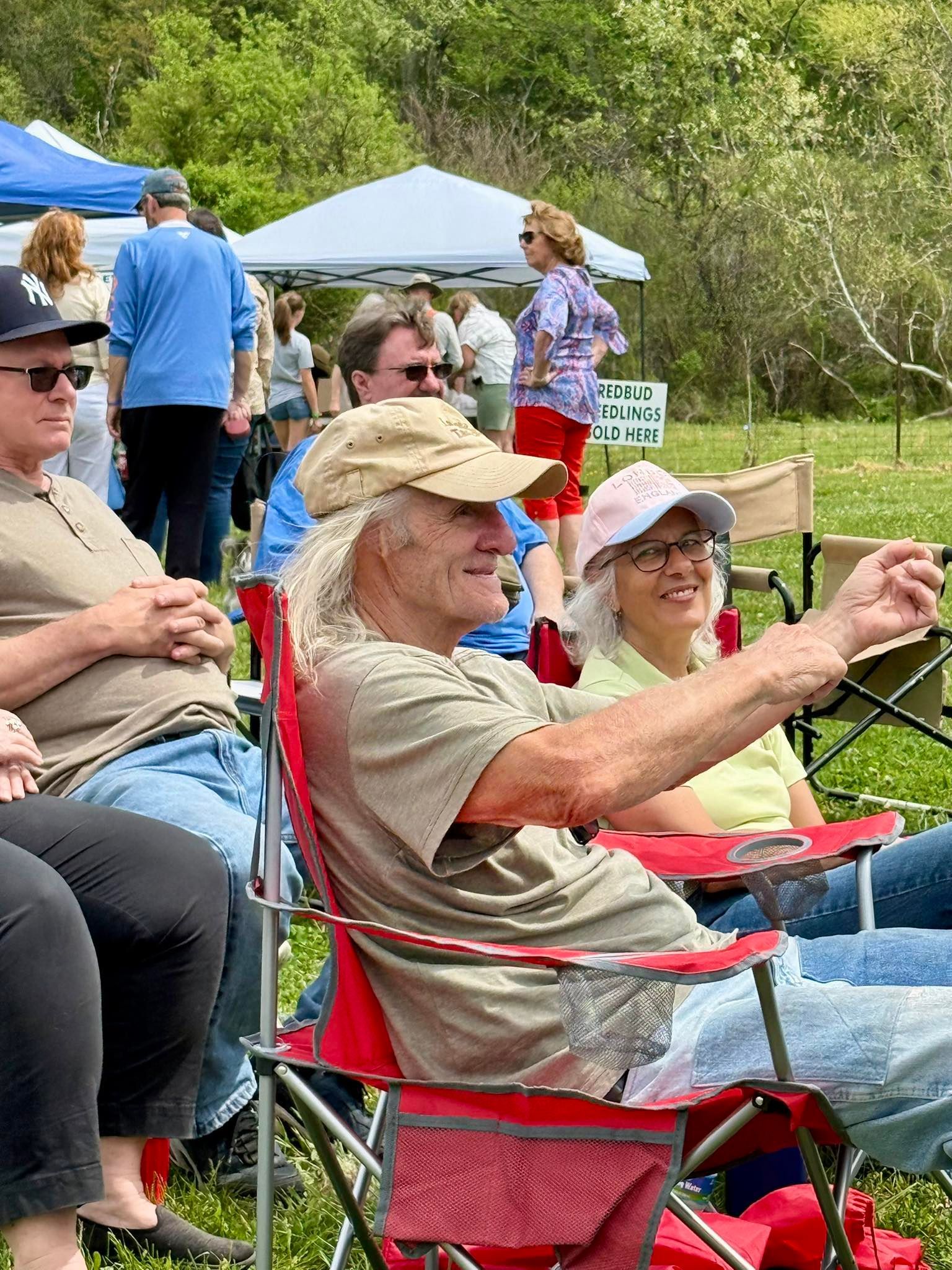 People seated in folding chairs at an outdoor gathering, one person taking a photo with a smartphone.
