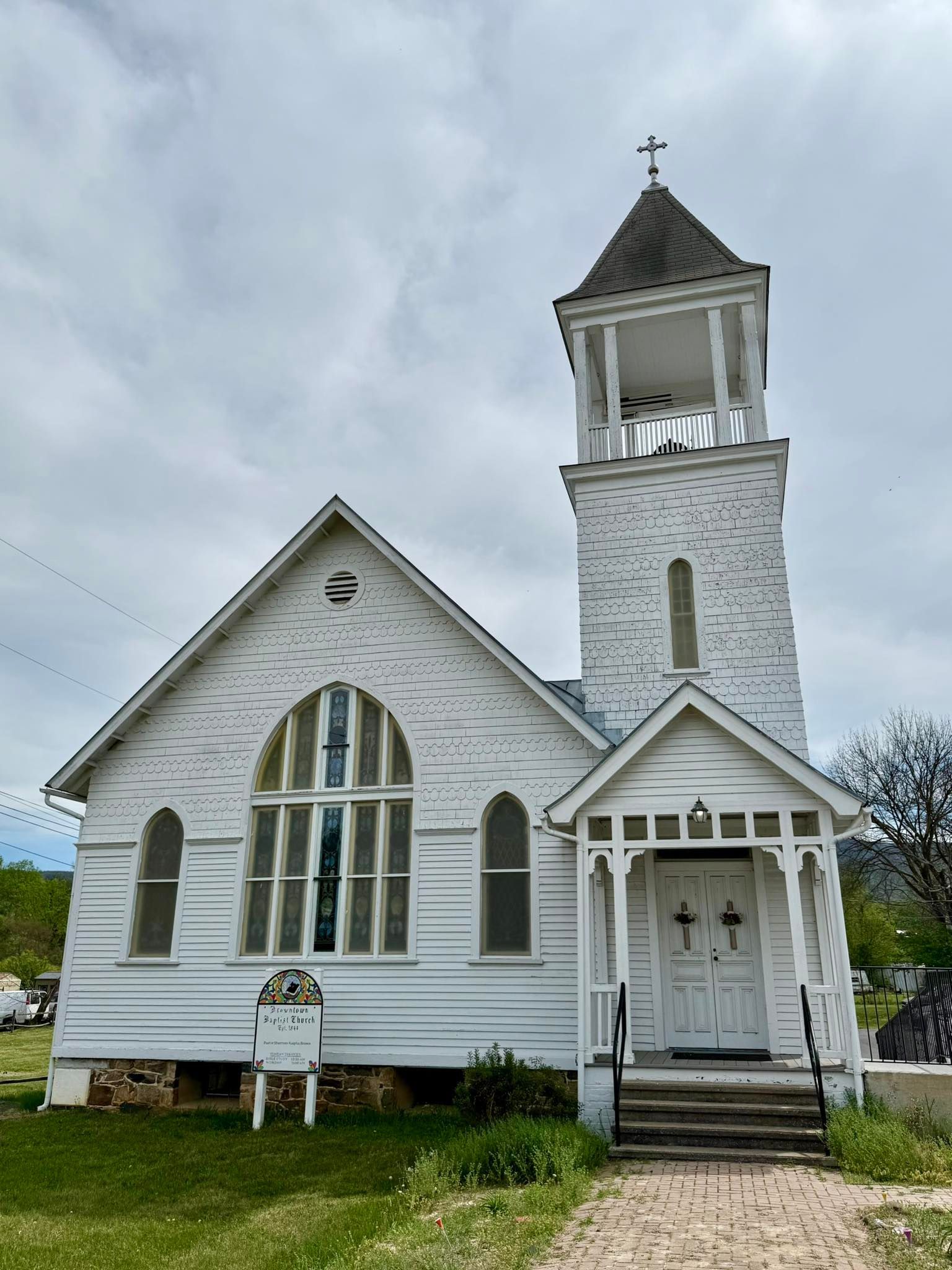 Small white wooden church with a tall steeple, arched windows, and a front porch on a grassy lot.