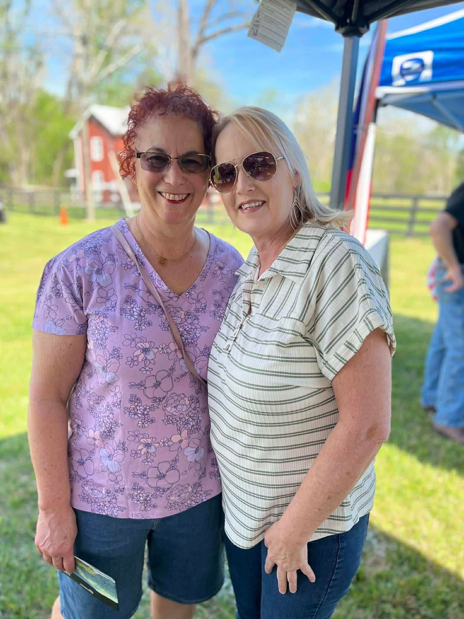 Two smiling people standing together outdoors on a sunny day, wearing sunglasses and casual summer clothes