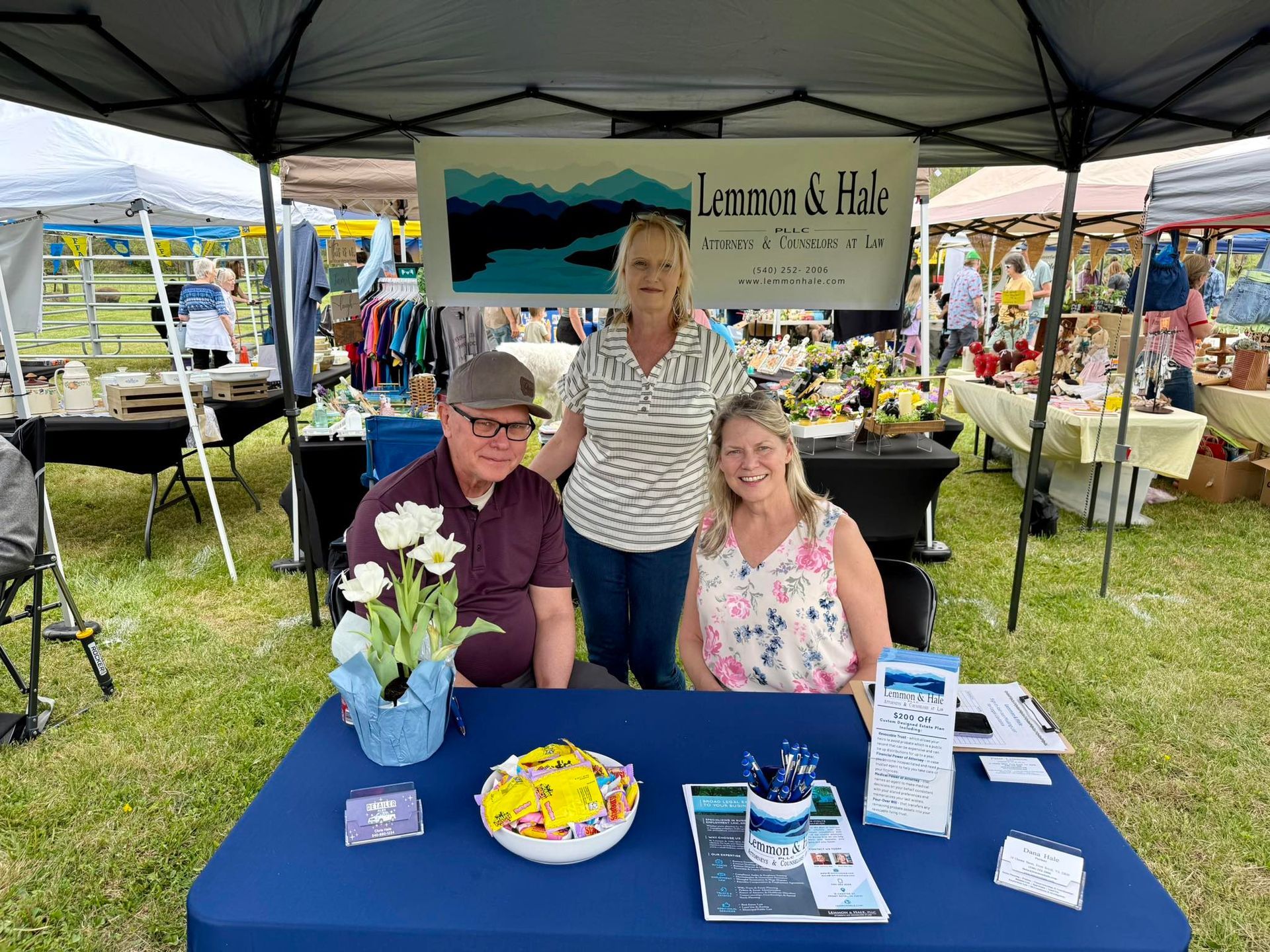Three people at a vendor booth with a blue tablecloth, flowers, and brochures under a canopy at a market.