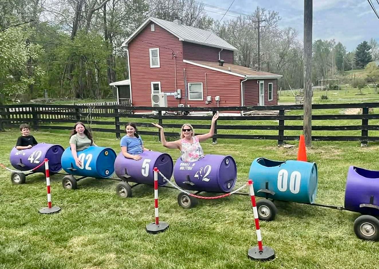 Four people ride colorful barrel train carts past a red barn on a grassy farm