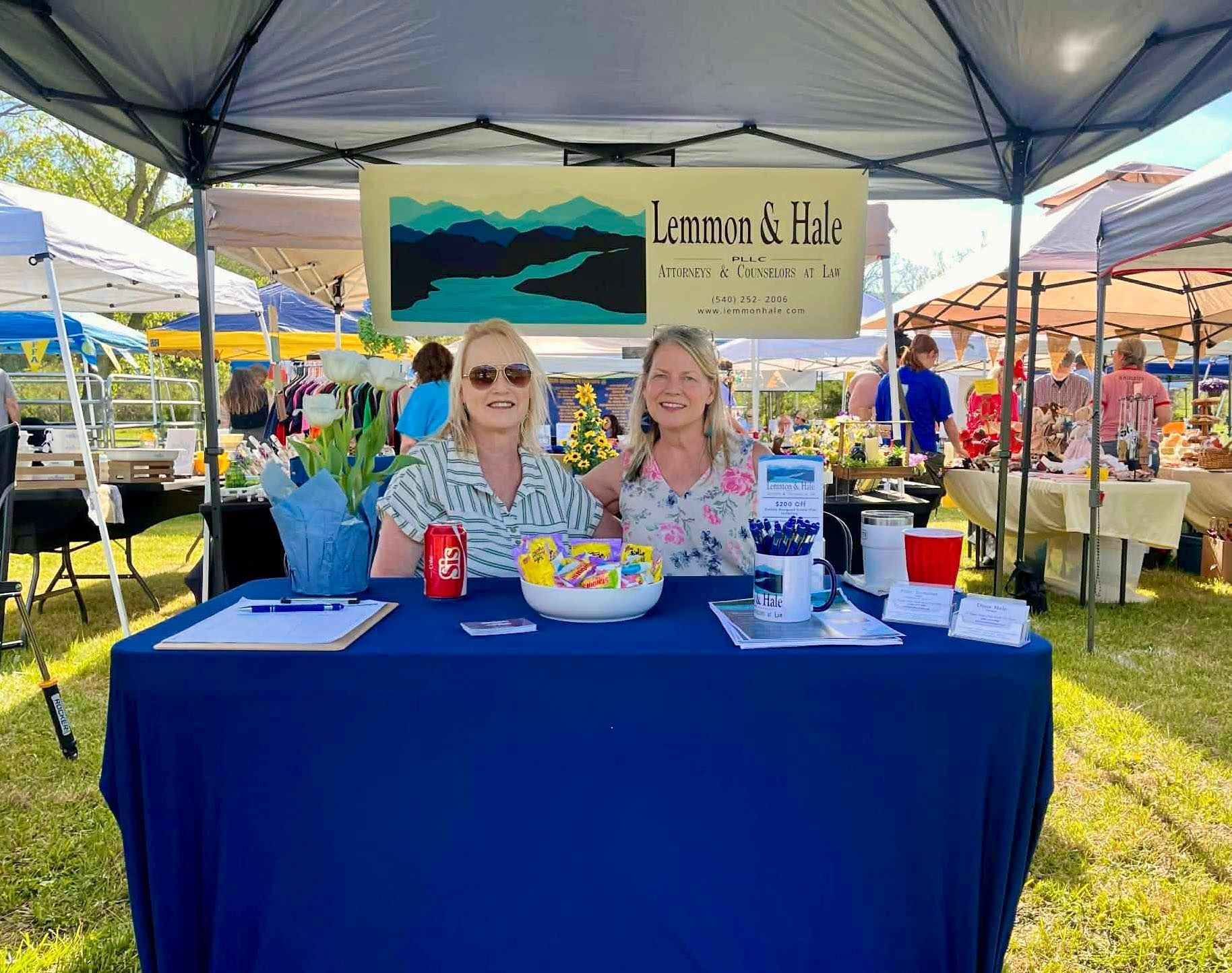 Two women behind a blue table at a lemon stand under a tent, with a colorful “Lemon & Lila” sign.