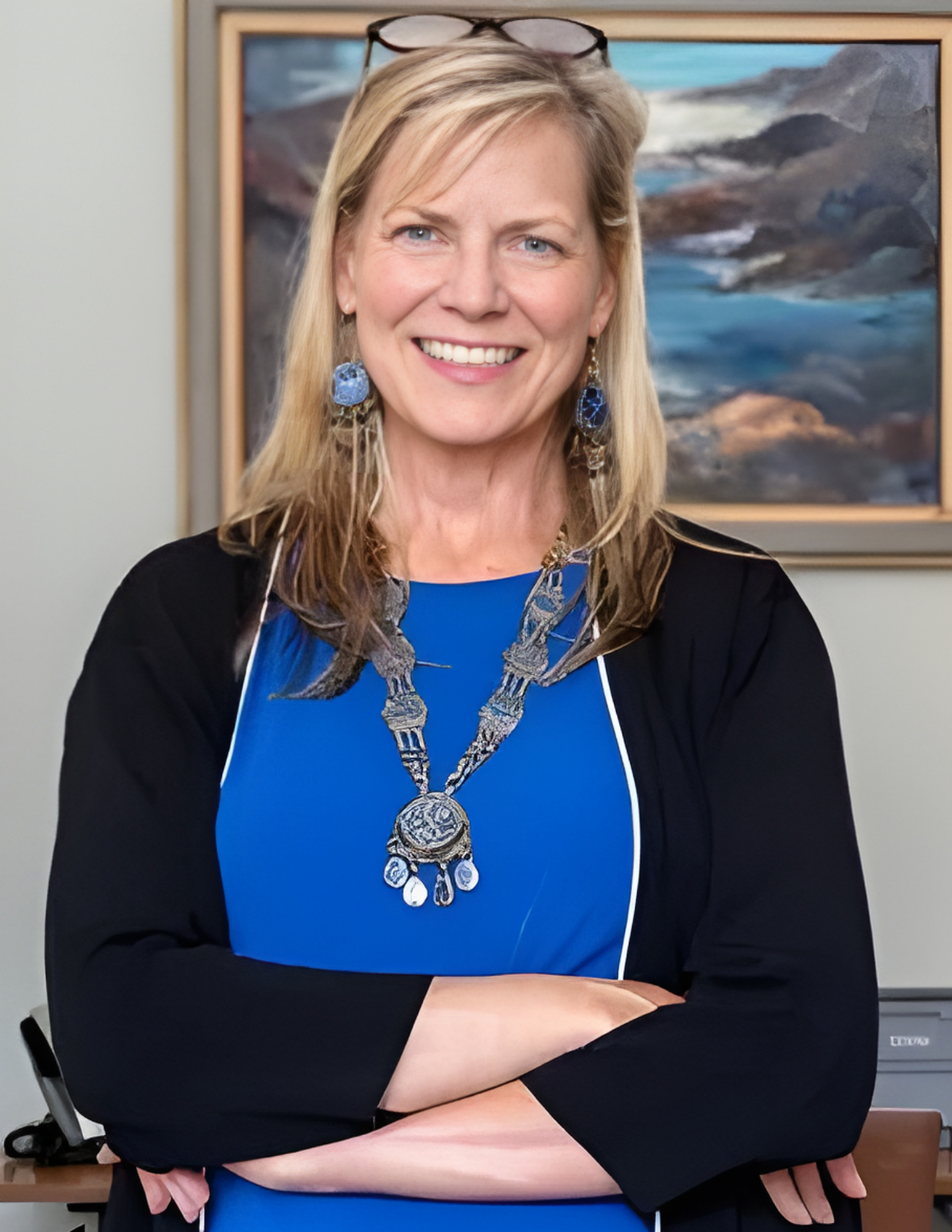 A smiling woman with blonde hair, wearing a blue top and layered silver necklace, stands with arms crossed in an office.