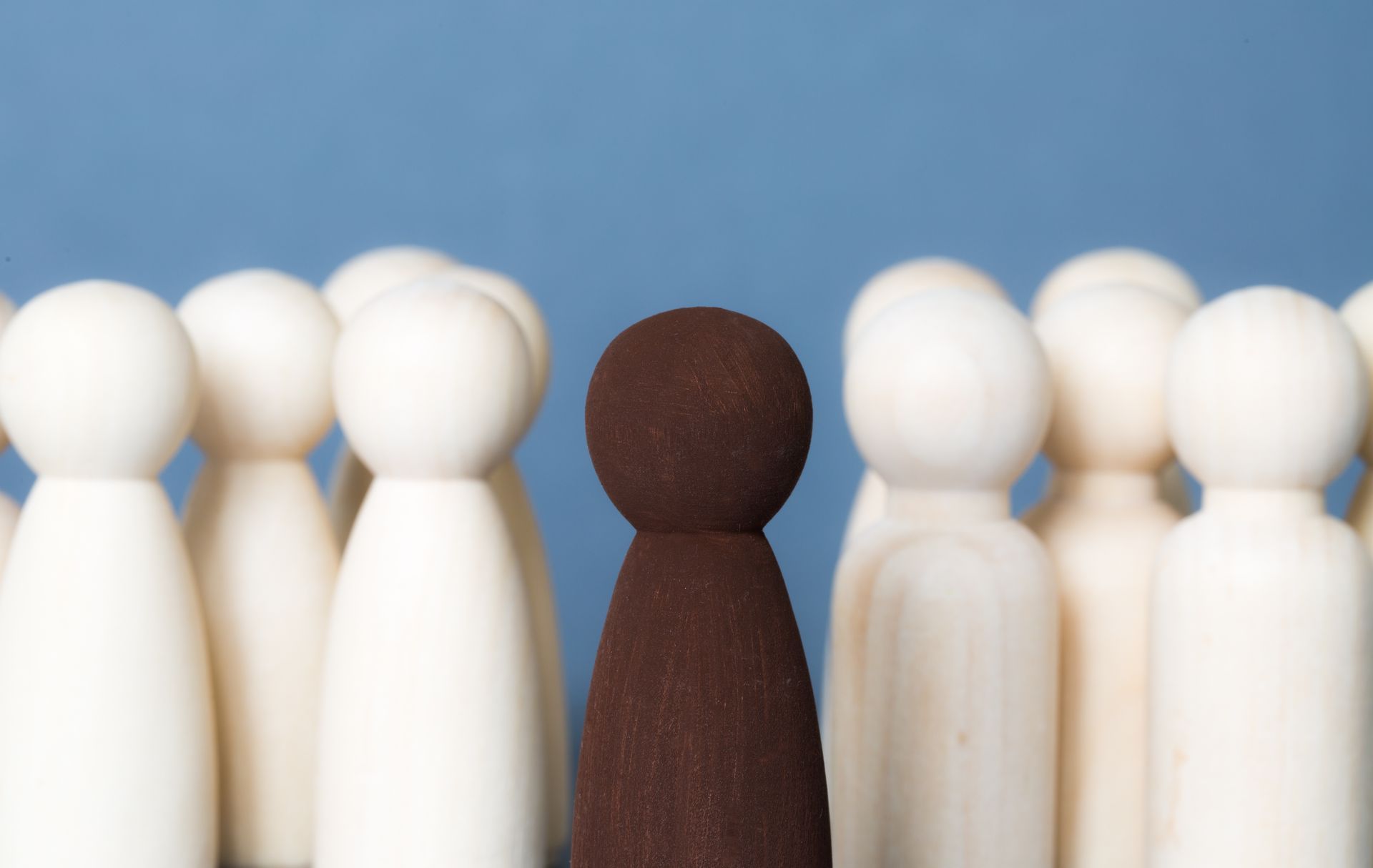A dark brown wooden peg doll stands in the center of a group of light wooden peg dolls against a blue background.