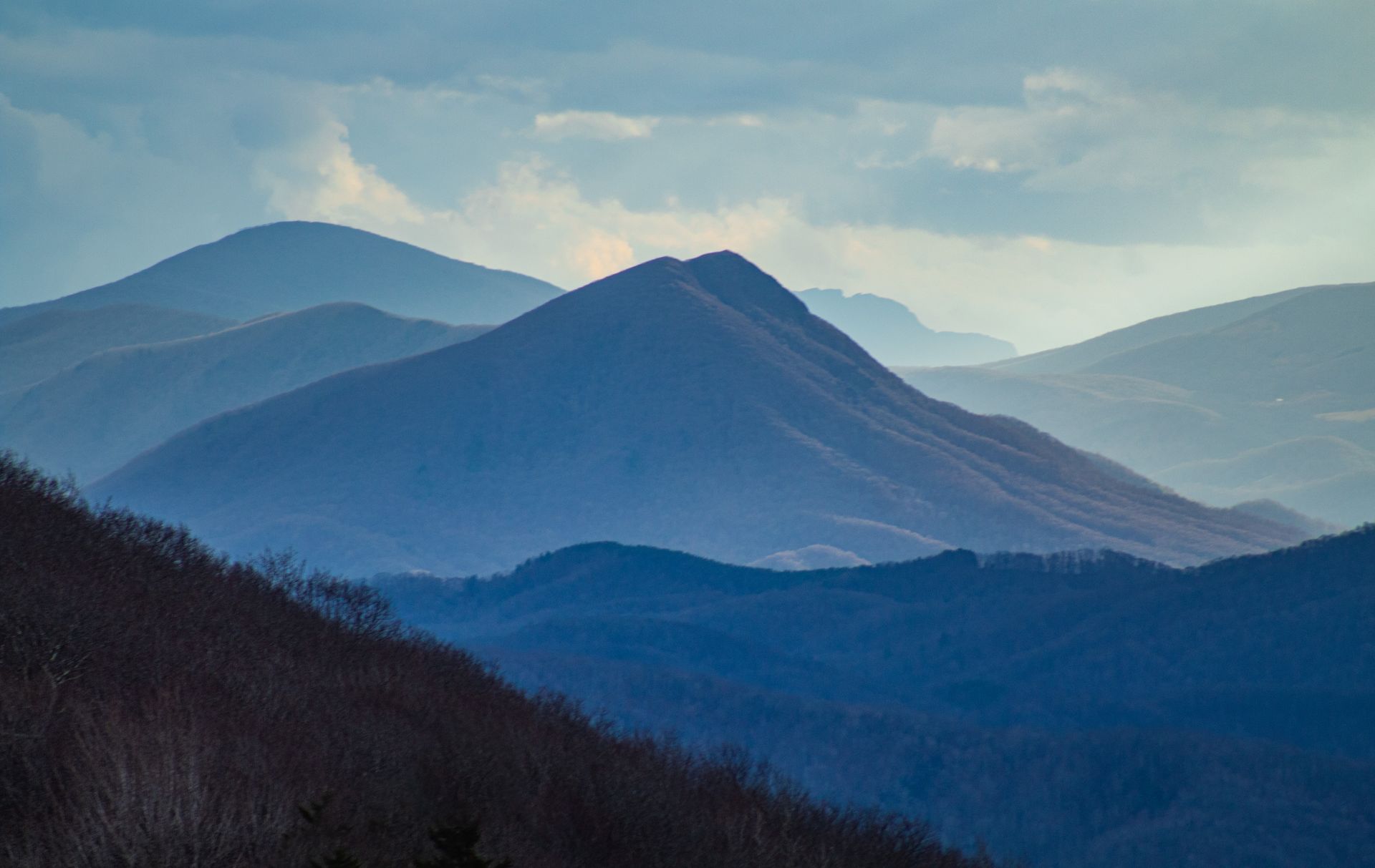 Layers of mountains fade into the distance under a cloudy sky, showcasing shades of blue and hazy silhouettes.