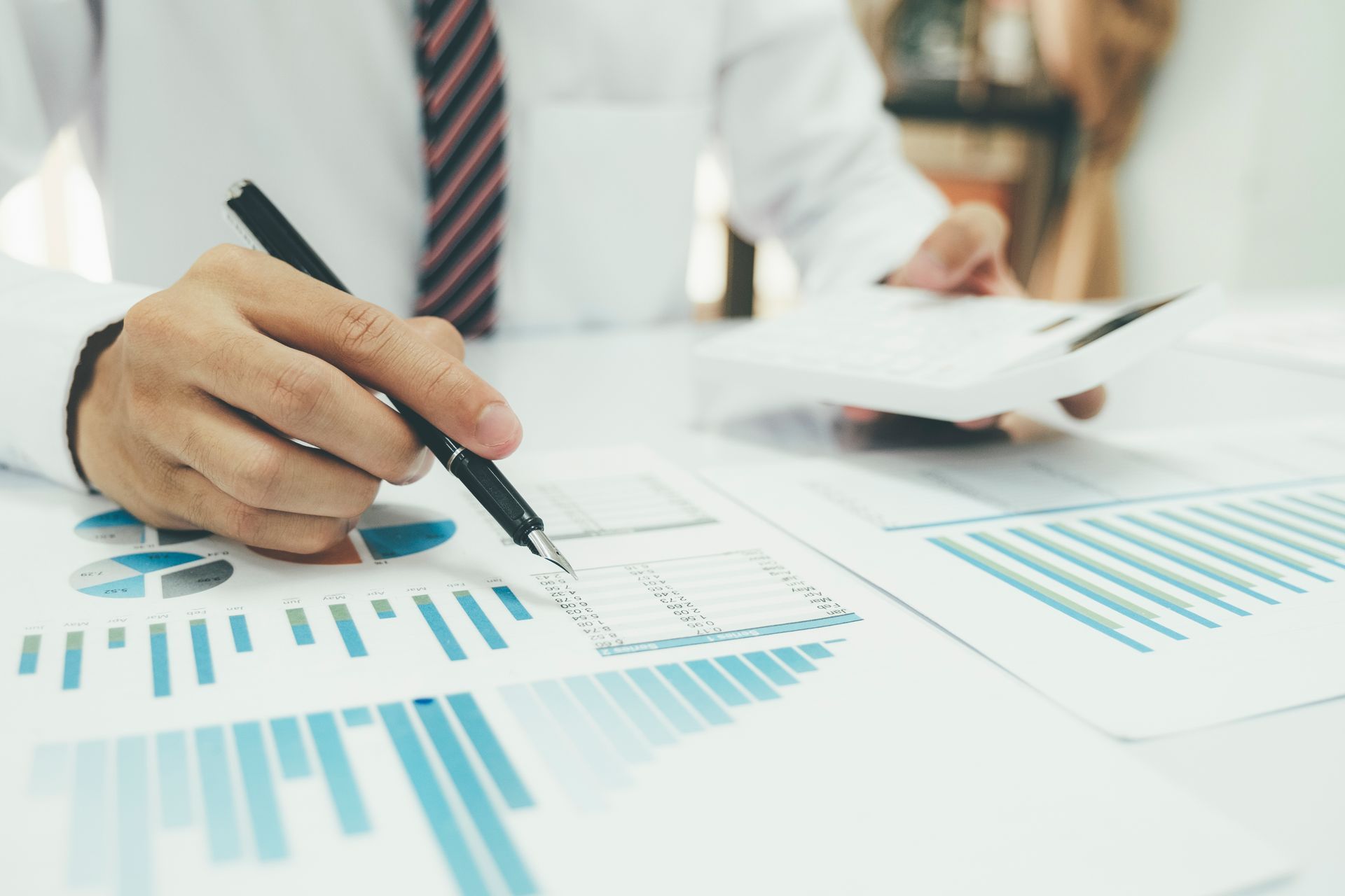 A person in a white dress shirt and tie uses a pen to review financial charts on a desk with a calculator nearby.