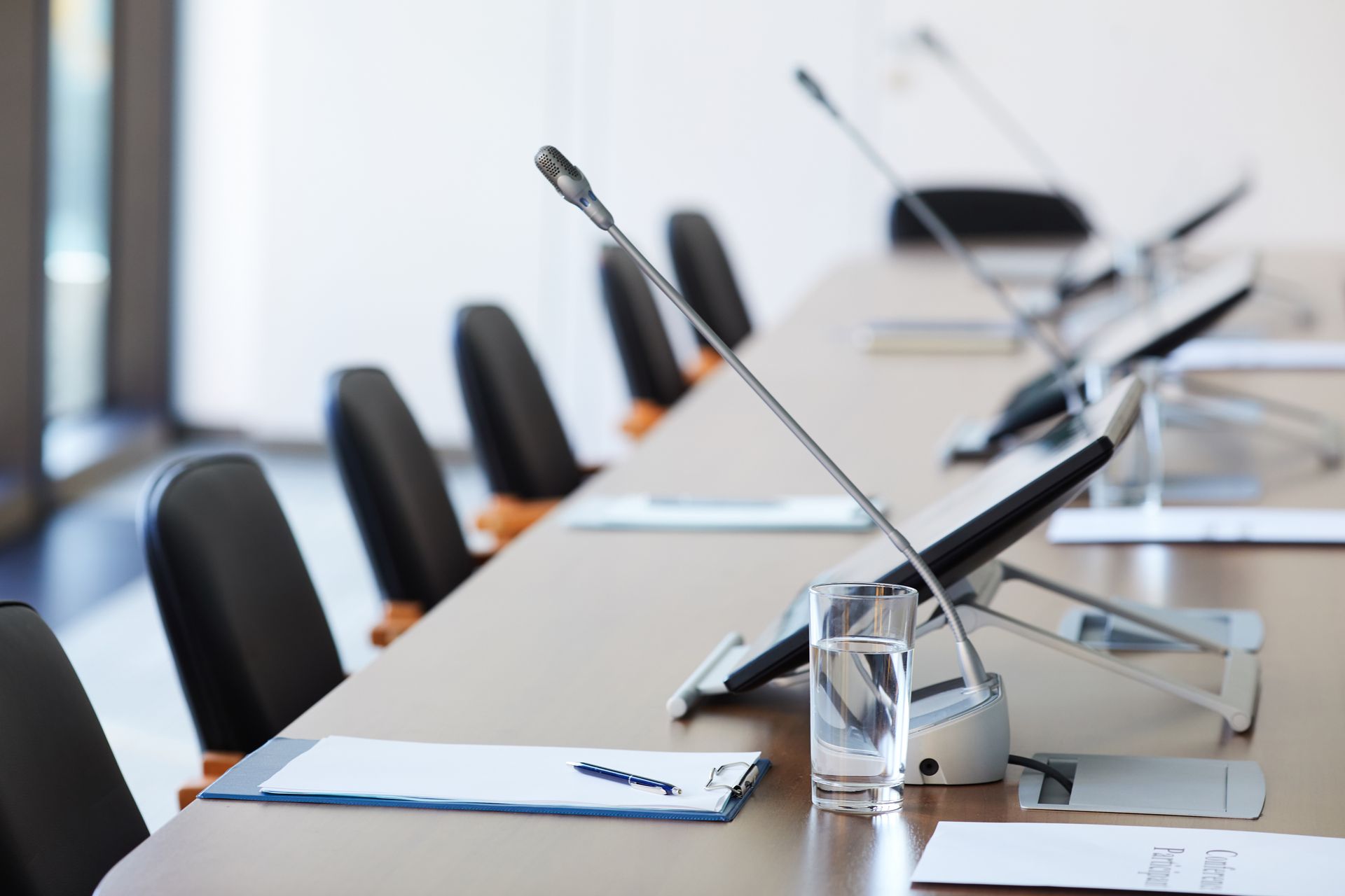 A conference table lined with black chairs, microphones, tablet stands, and water glasses in a brightly lit room.