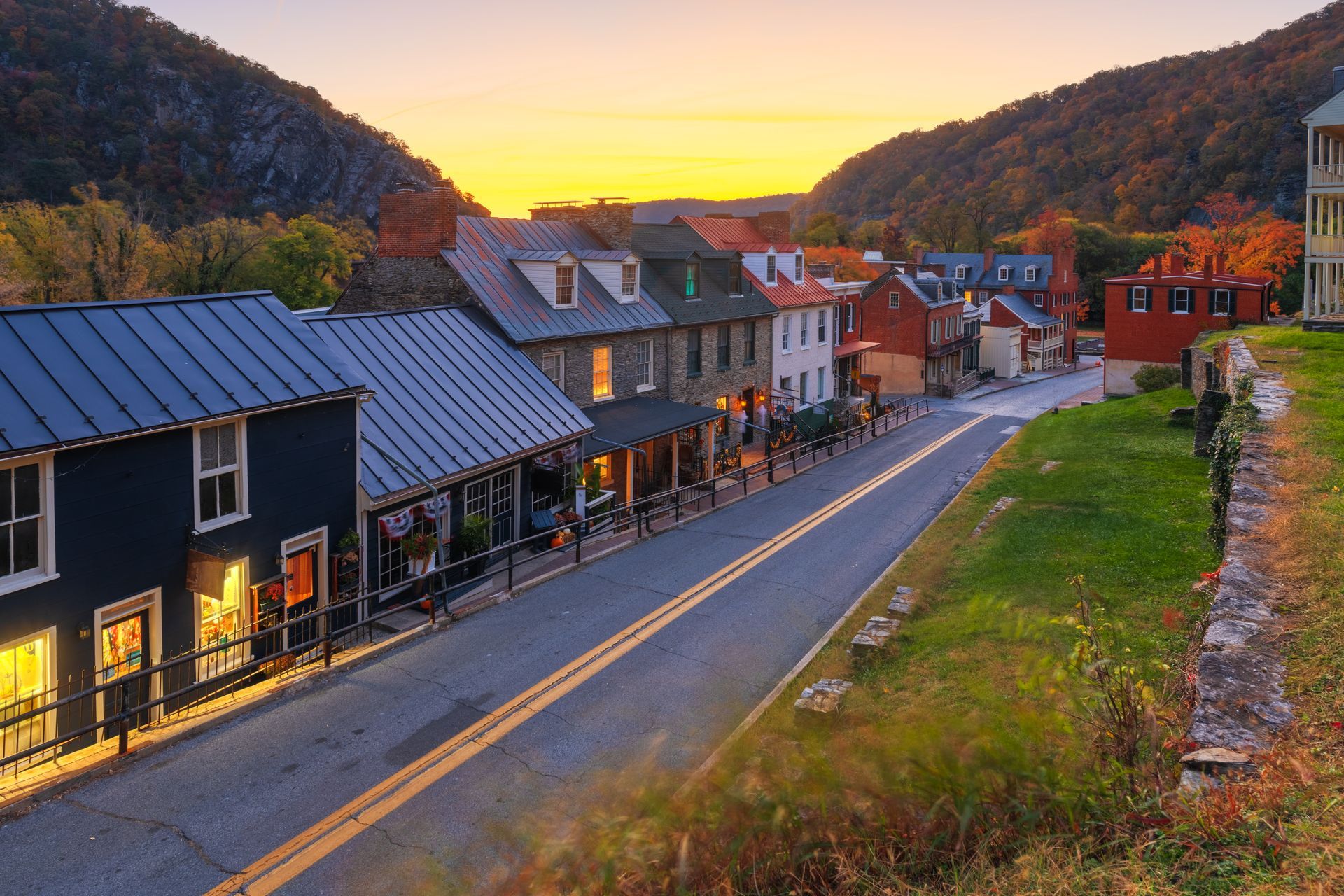 A historic street with stone buildings at dusk, nestled between mountains under a vibrant yellow and orange sky.