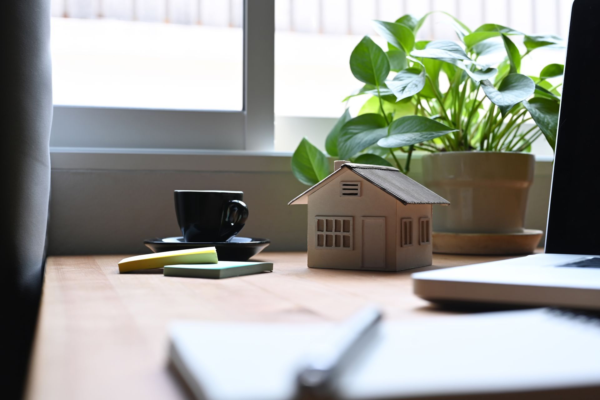A wooden model house, a coffee cup, and a plant on a desk near a window with a notebook and laptop in the foreground.