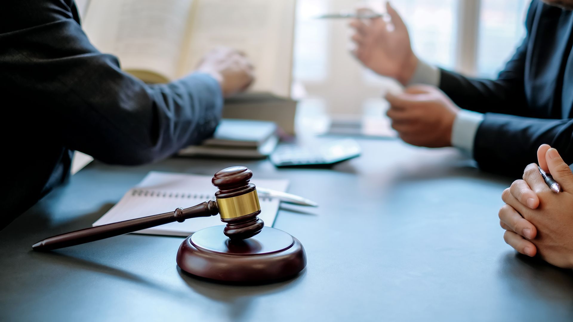 A wooden gavel rests on a desk in a boardroom setting while two individuals in suits consult documents and converse.