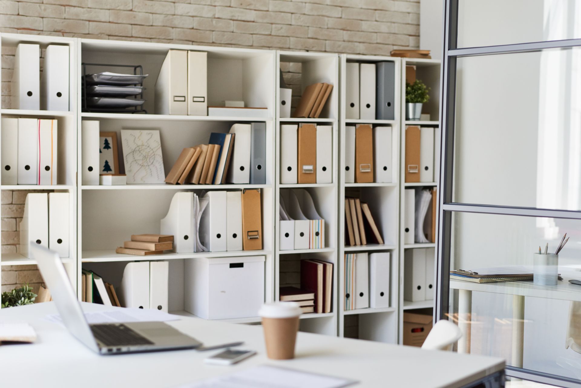 A well-organized office with white bookshelves filled with binders and files, and a laptop on a white desk in the foreground.