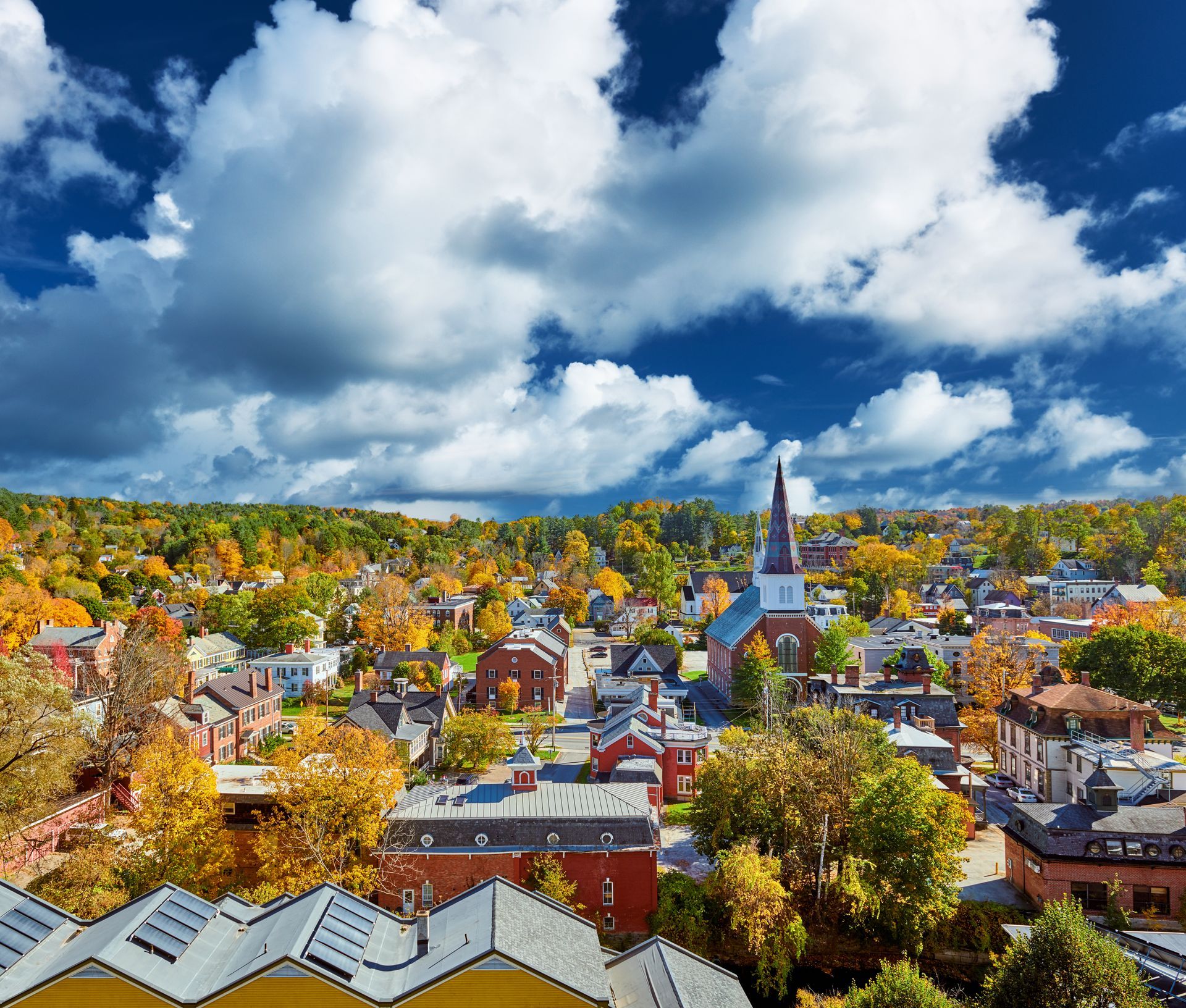 A scenic high-angle view of a small town nestled among vibrant autumn foliage under a bright blue sky with large clouds.