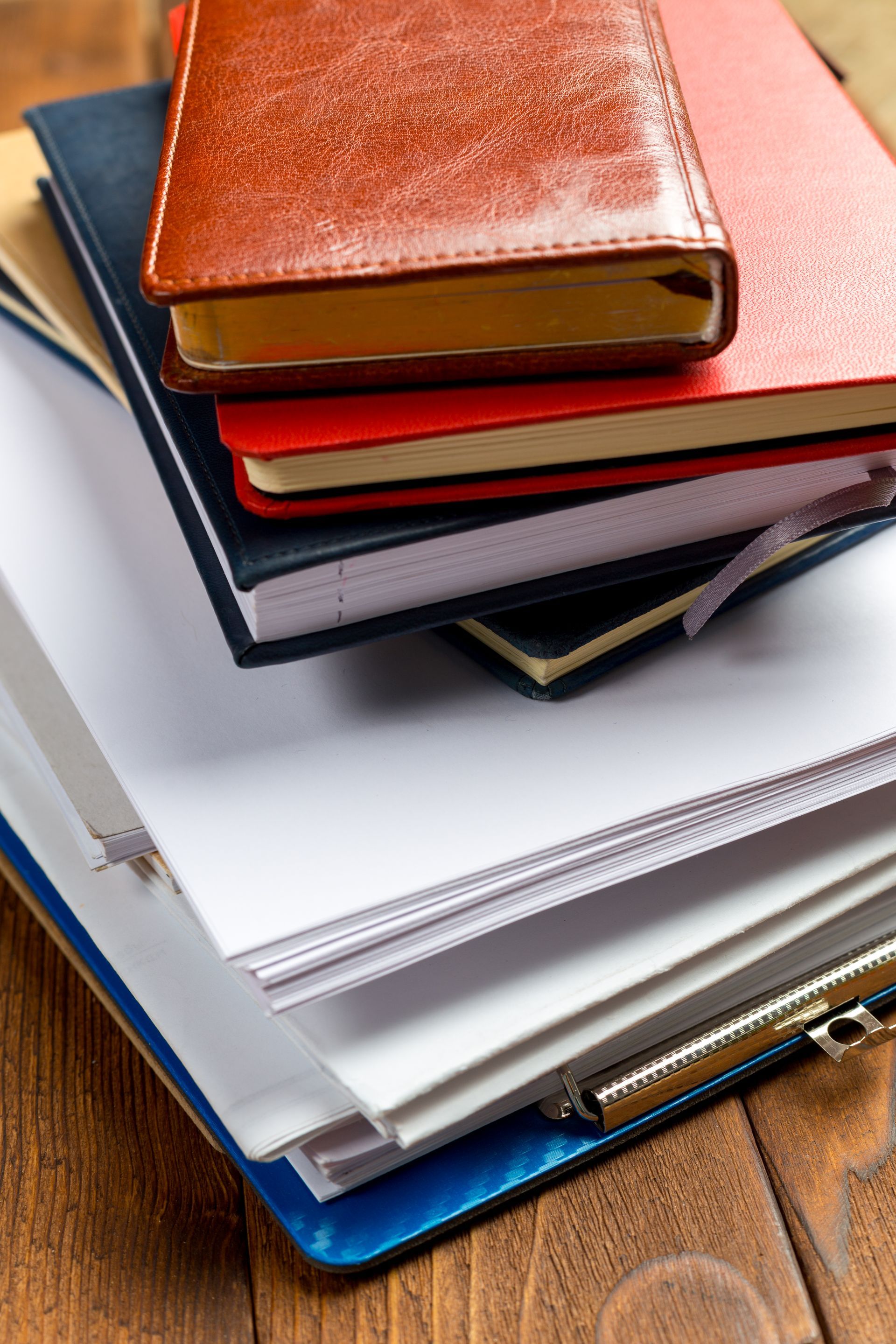 A stack of various notebooks and loose papers resting on a wooden surface.