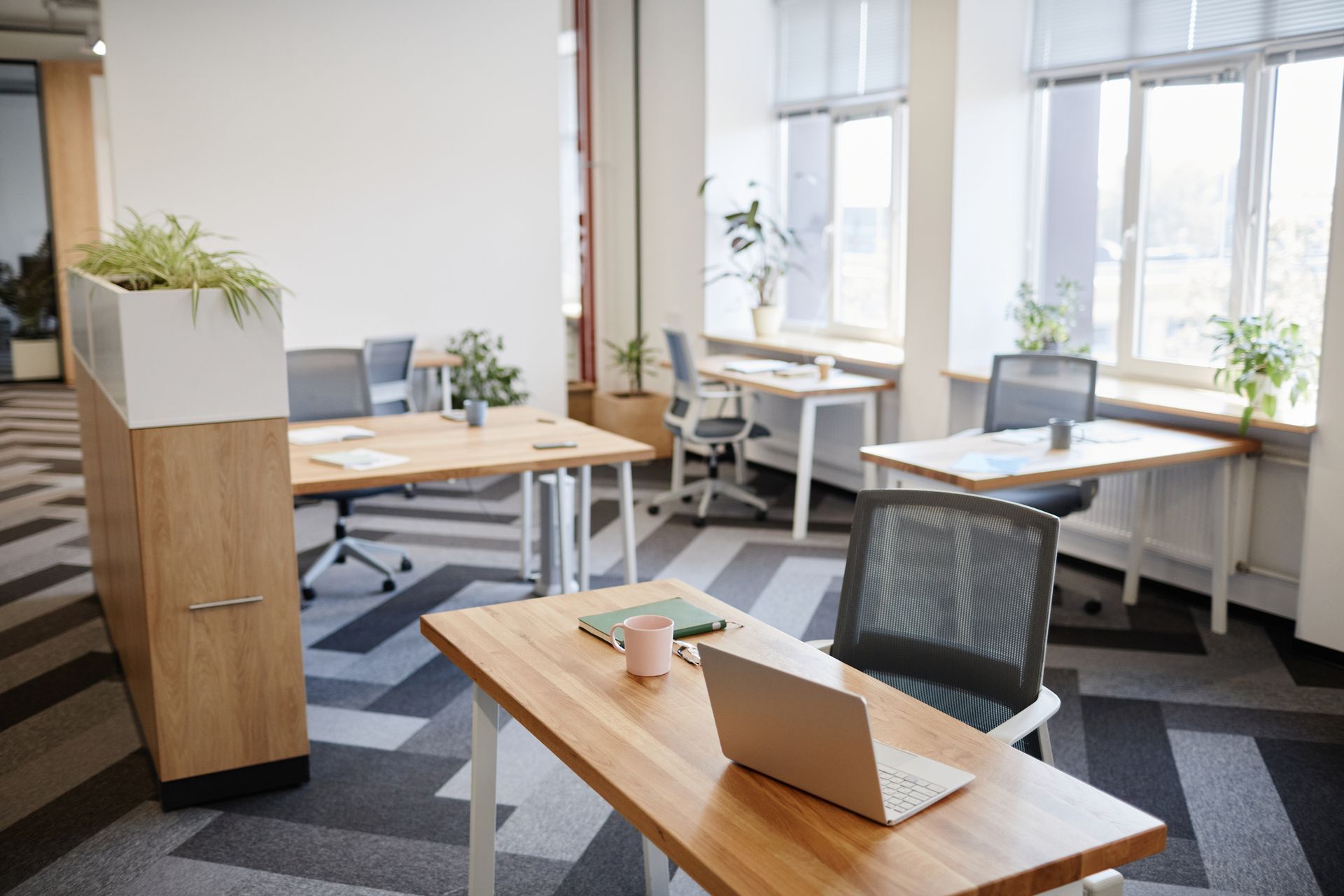 A bright office space with multiple wooden desks, ergonomic chairs, and large windows, featuring a patterned floor.