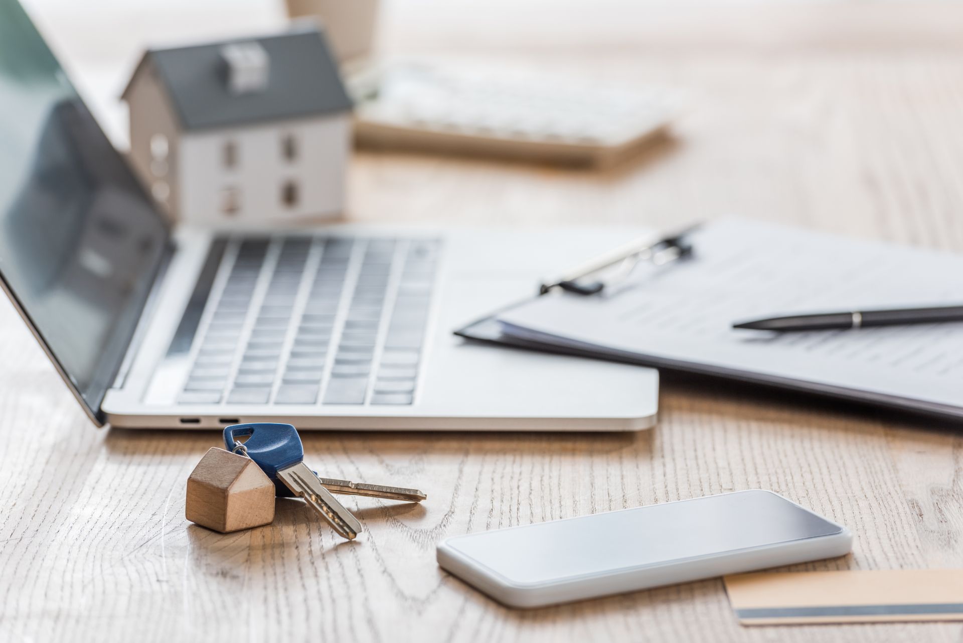 A laptop, clipboard with a pen, keys with a house charm, and a smartphone on a wooden desk.