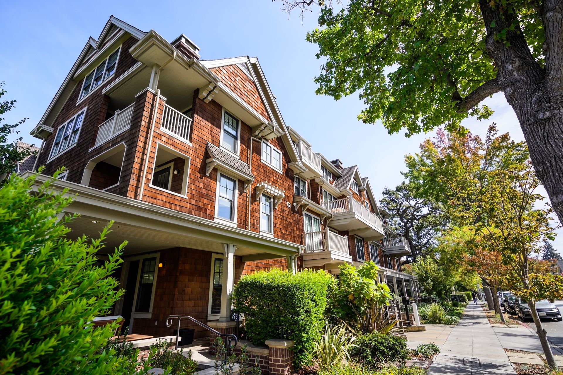 A multi-story building with brown shingle siding, balconies, and a covered porch, set on a sunny street with trees.