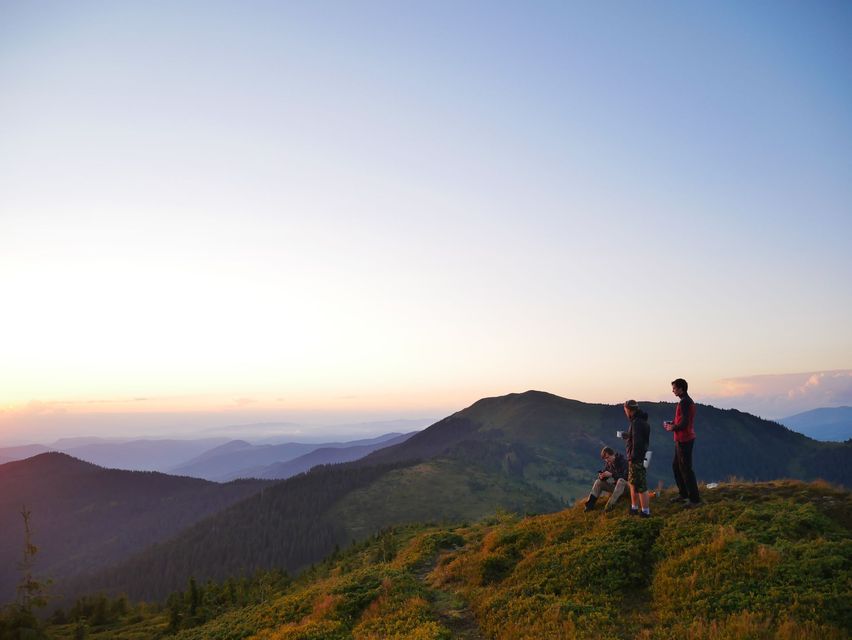 Three people stand on a grassy mountain ridge during sunrise, overlooking layers of distant mountain ranges.
