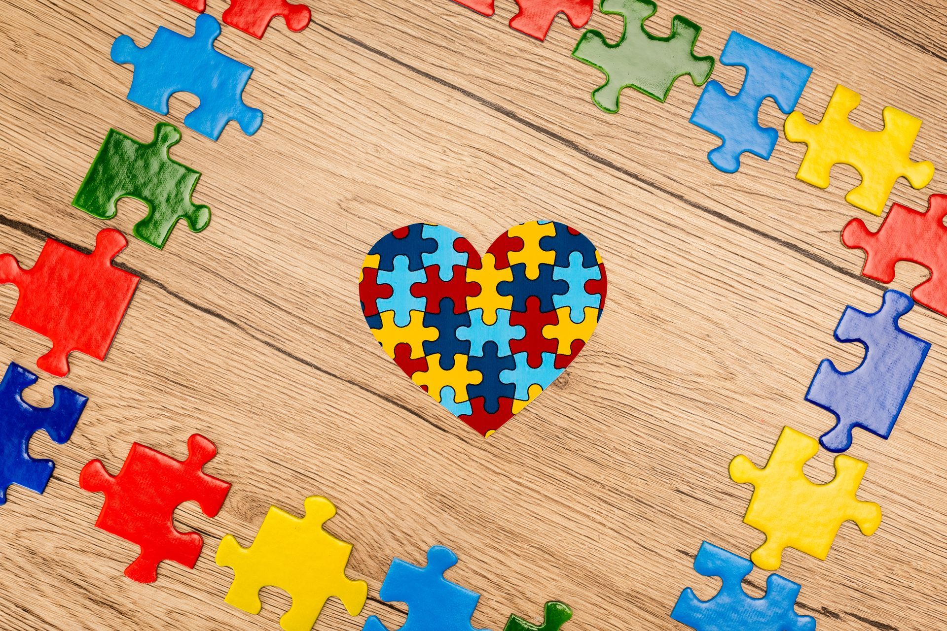 A heart made of colorful puzzle pieces sits in the center of a wooden surface, surrounded by loose, colorful puzzle pieces.