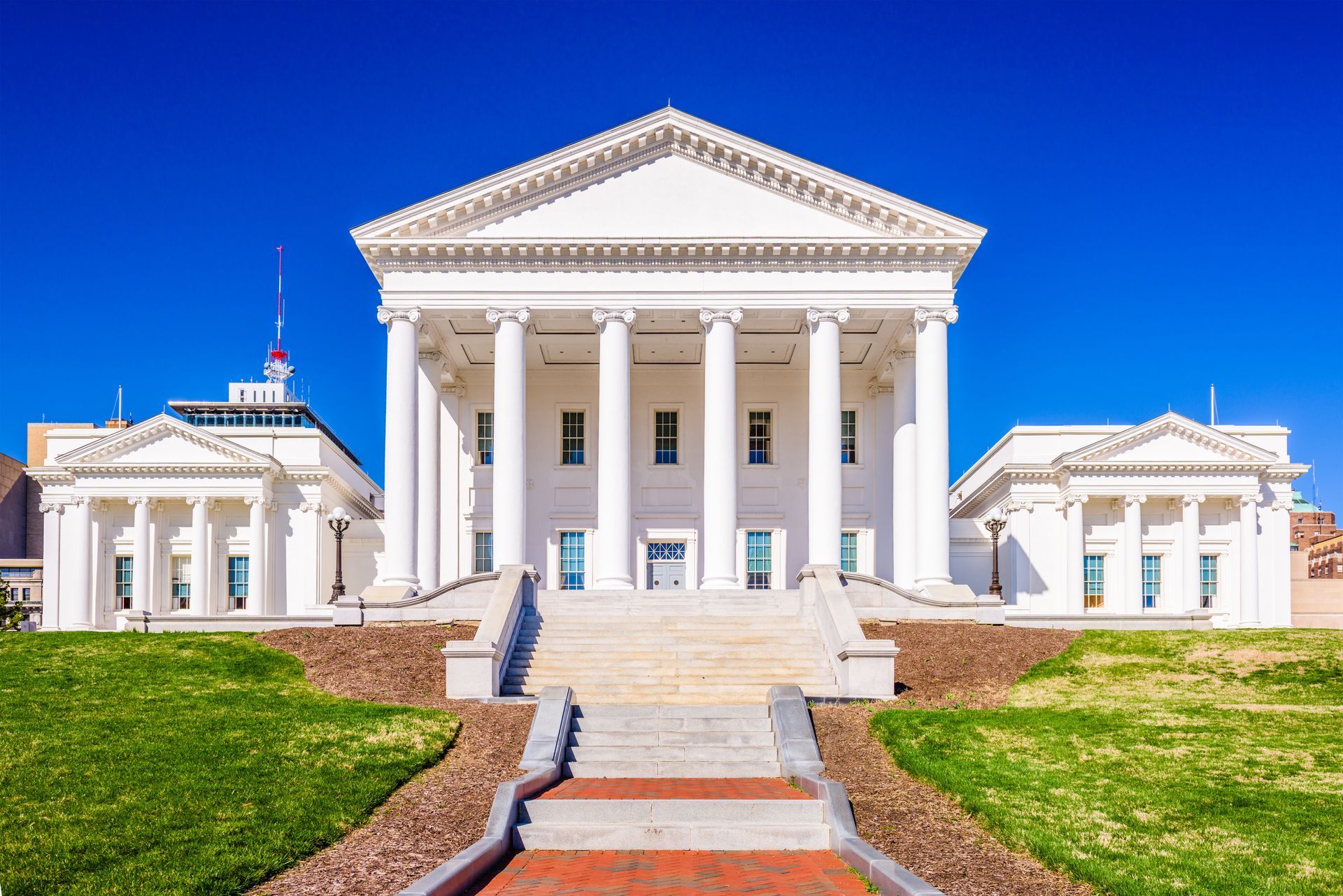 The Virginia State Capitol building with white neoclassical columns, a triangular pediment, and green lawns under a blue sky.