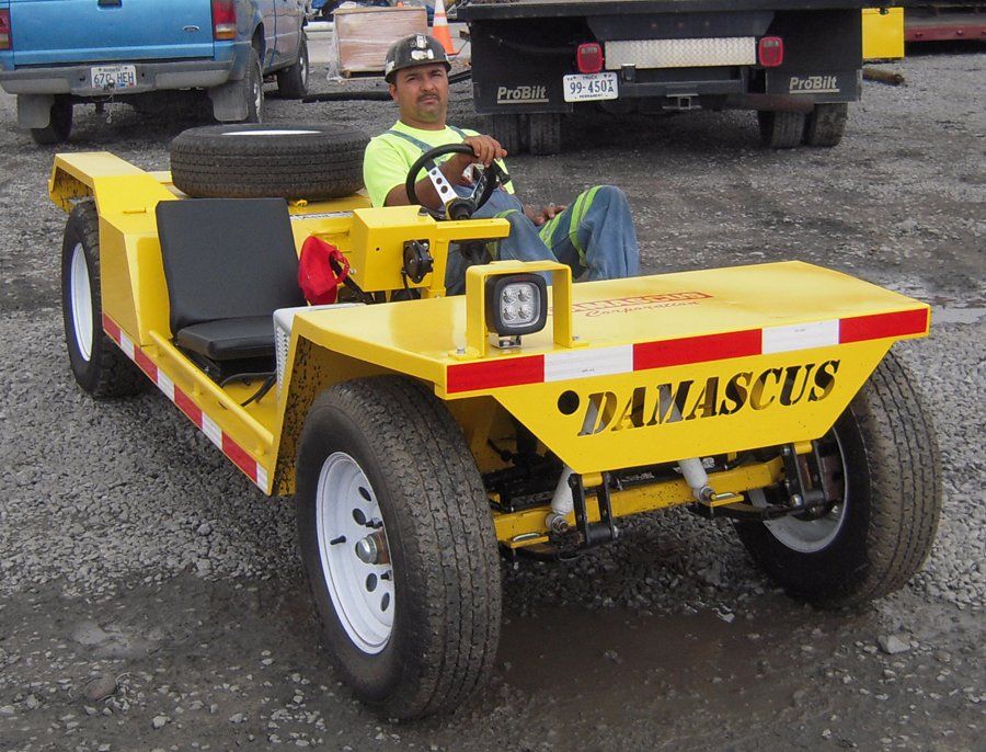 Man in a yellow industrial vehicle. He's driving and wearing a hard hat, sitting outdoors. The vehicle has 