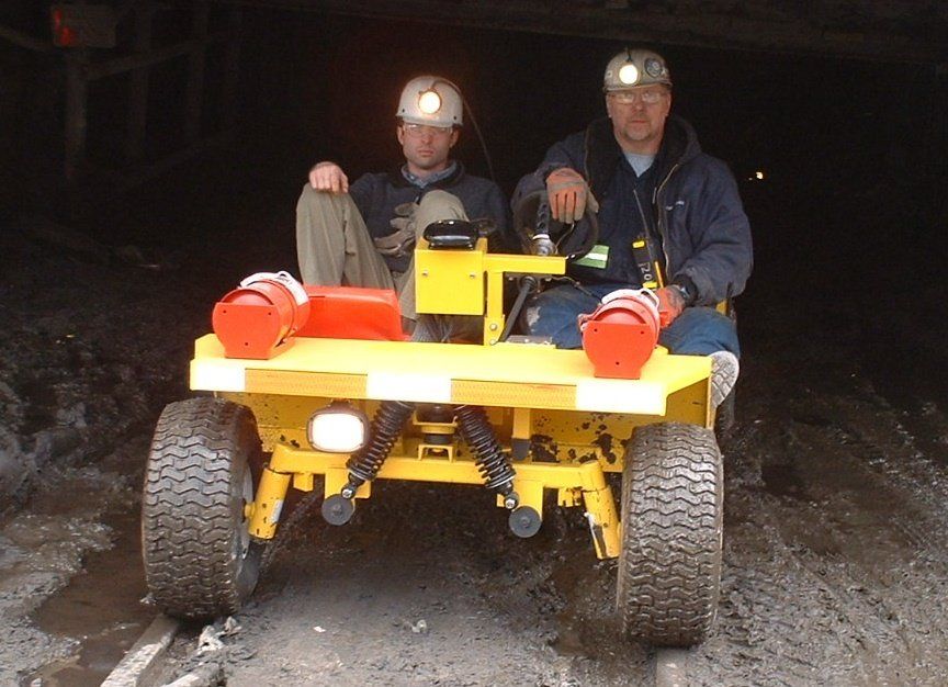 Two people in hard hats sit on a yellow vehicle in a dark tunnel, likely a mine.