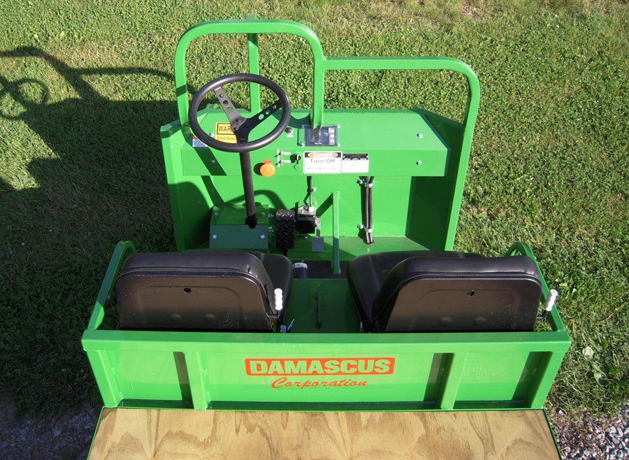 Green Damascus Corporation utility vehicle with two seats, steering wheel, and safety bar on wooden platform.