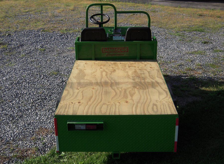 Green utility cart with plywood bed, black seats, and steering wheel, on gravel.