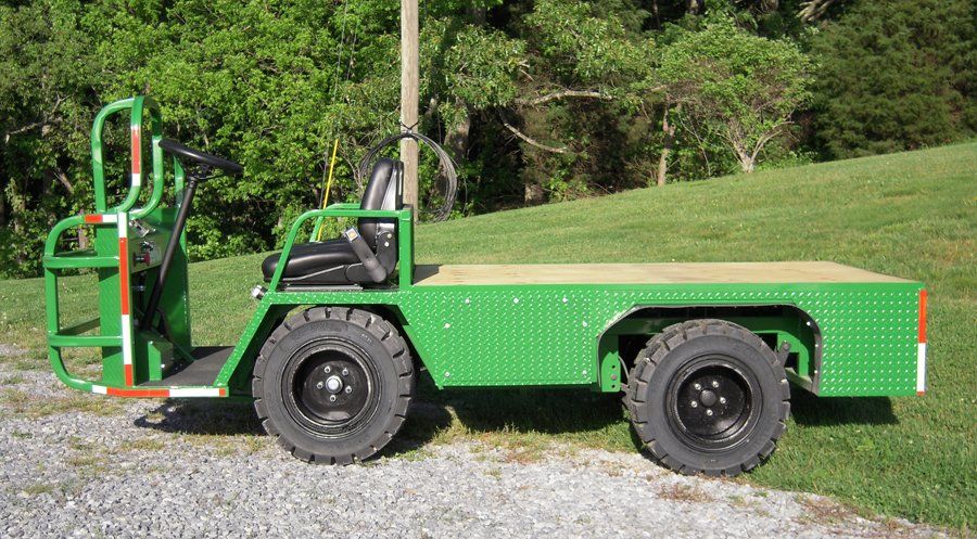 Green utility vehicle with a flatbed on gravel, next to grass.