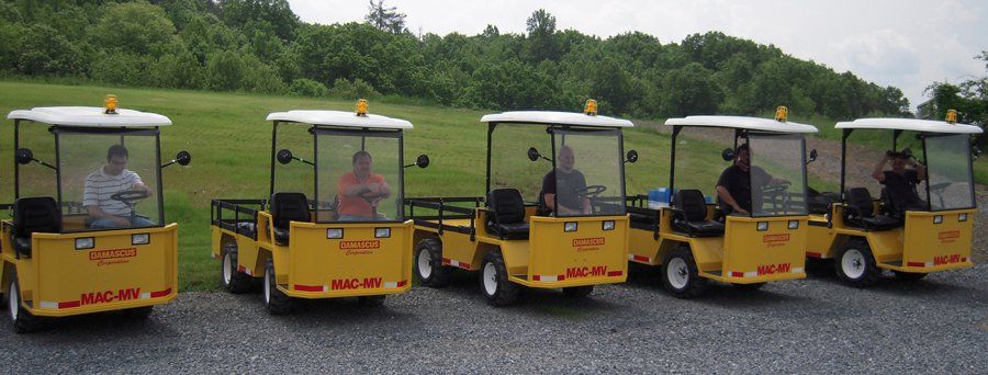 Five yellow utility vehicles with drivers sit in a row on gravel, with trees in the background.