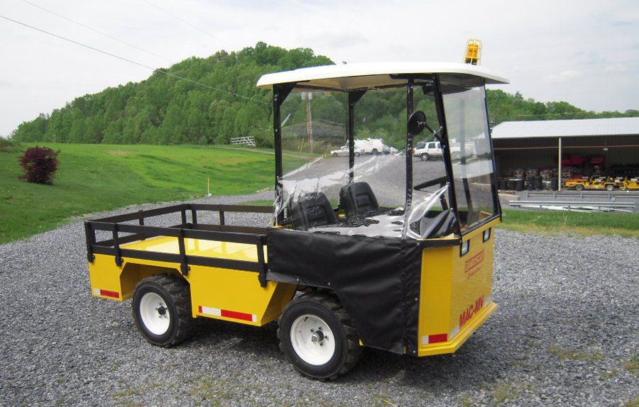 Yellow utility vehicle with a covered cab on gravel in front of a green hill.