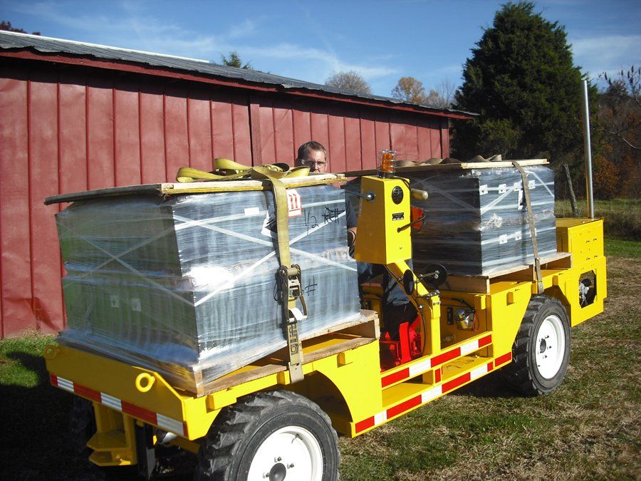 Yellow industrial vehicle carrying two wrapped pallets; person is in the driver's seat. Red barn in background.