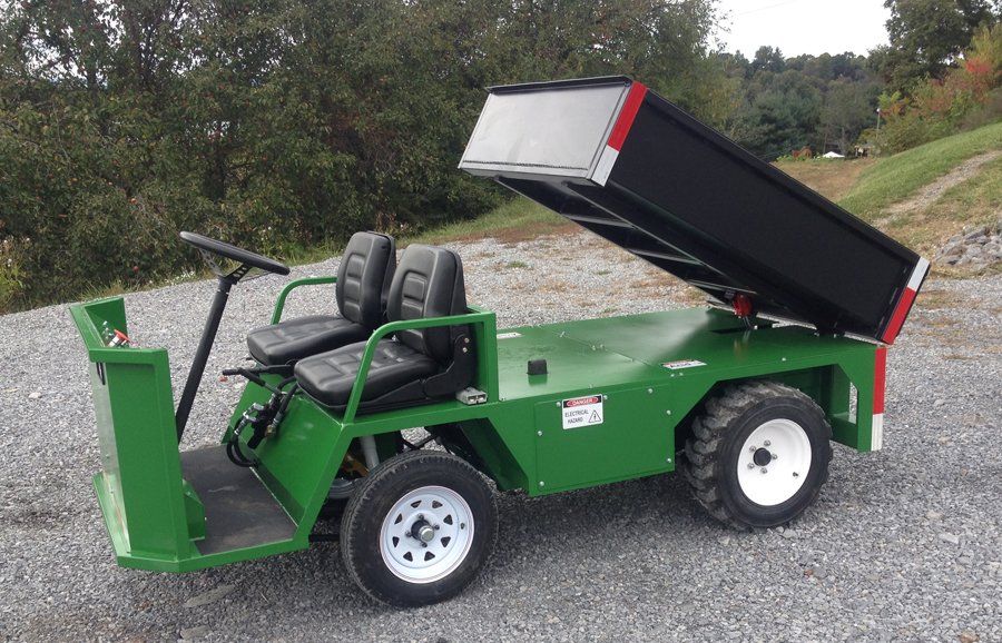 Green utility cart with raised black dump bed. Two seats, white wheels, outdoor setting.